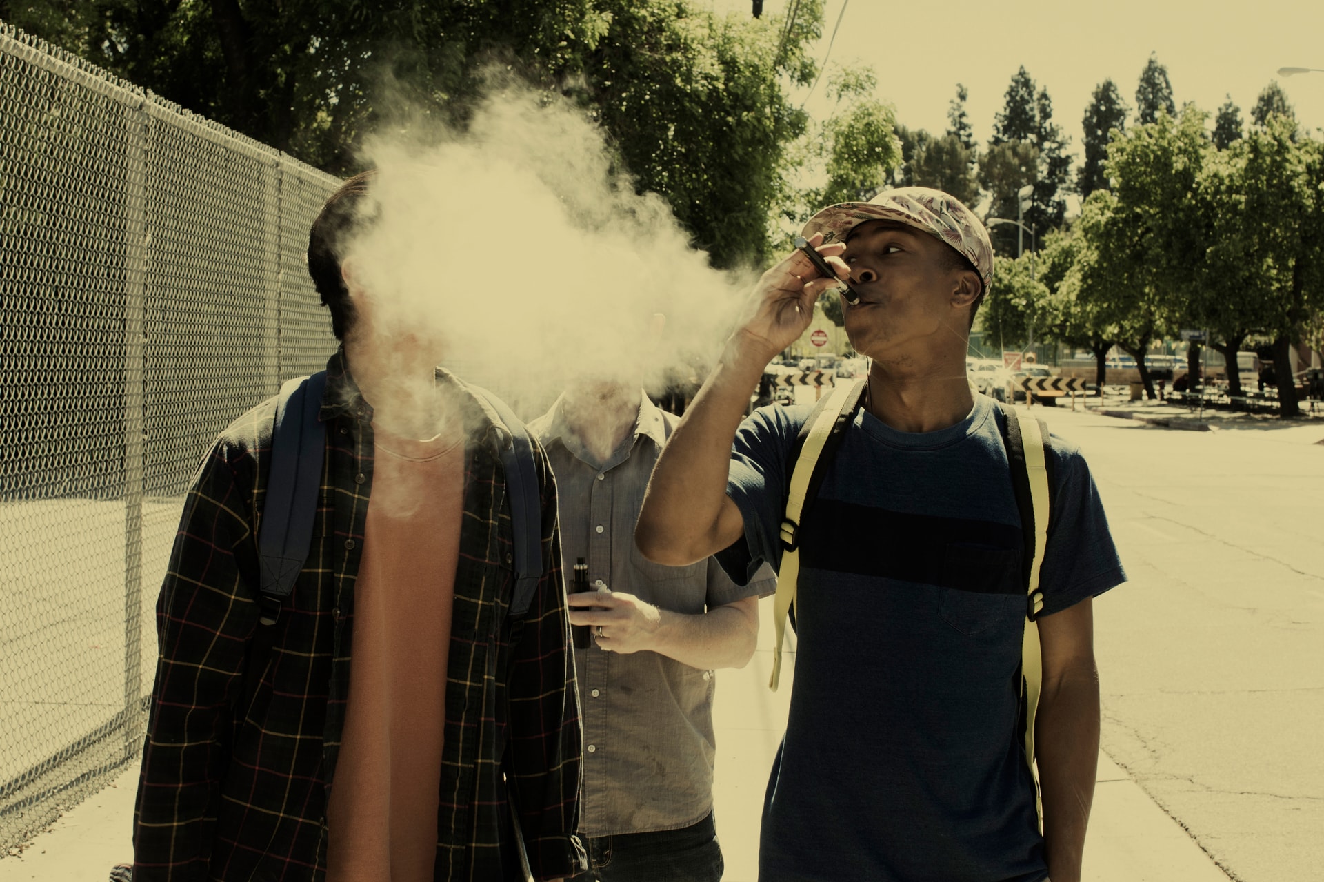 Three teenagers on a street, with one exhaling a cloud of secondhand vape into the others' faces
