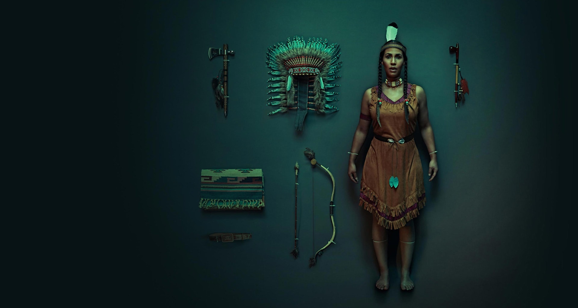 An American Indian woman dressed in traditional clothing standing in a dark room with American Indian accessories on display