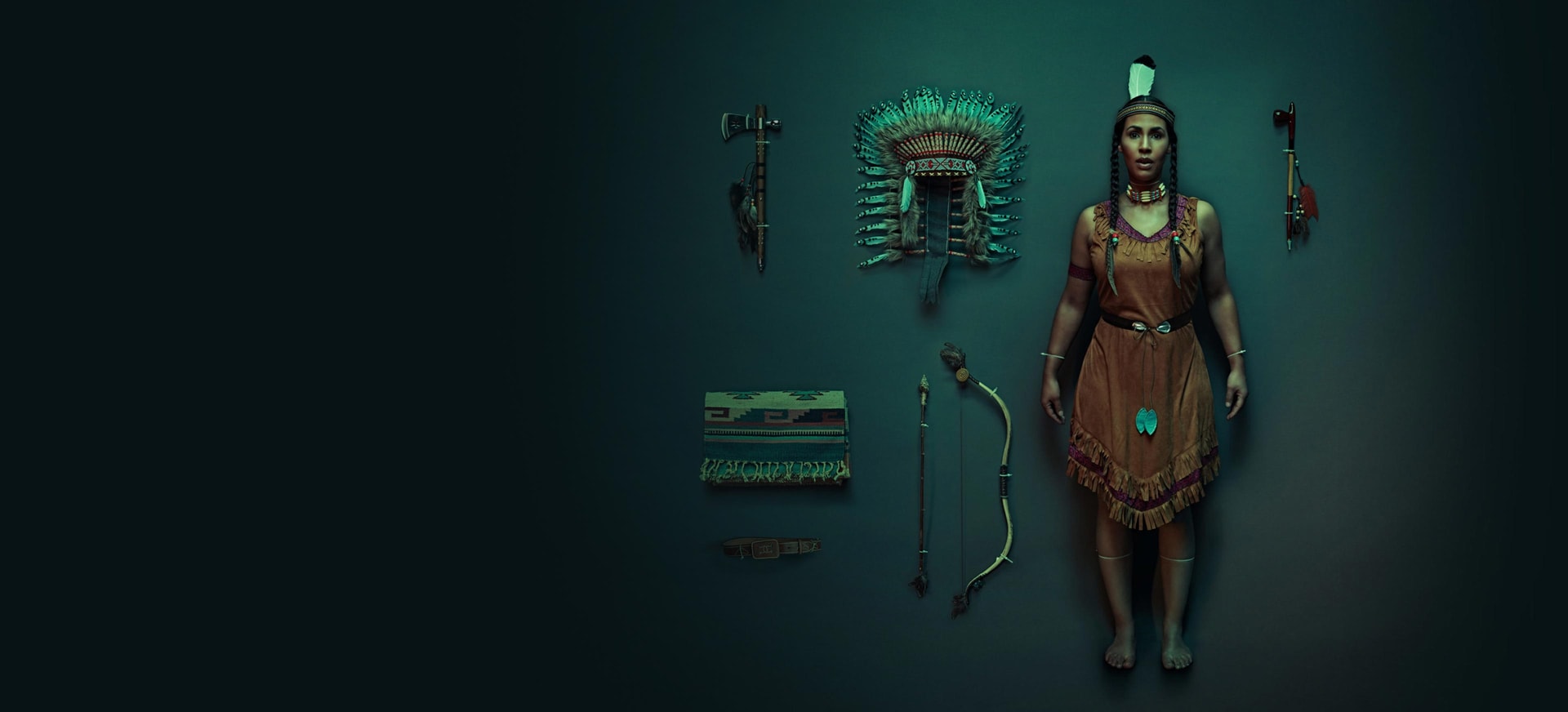 An American Indian woman dressed in traditional clothing standing in a dark room with American Indian accessories on display