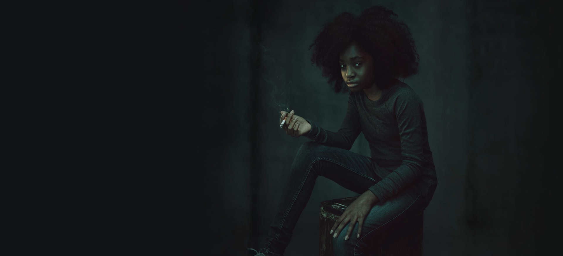A young African American/Black teen girl sitting on a stool in a dark room holding a cigarette