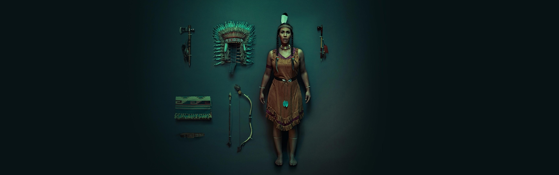 An American Indian woman dressed in traditional clothing standing in a dark room with American Indian accessories on display