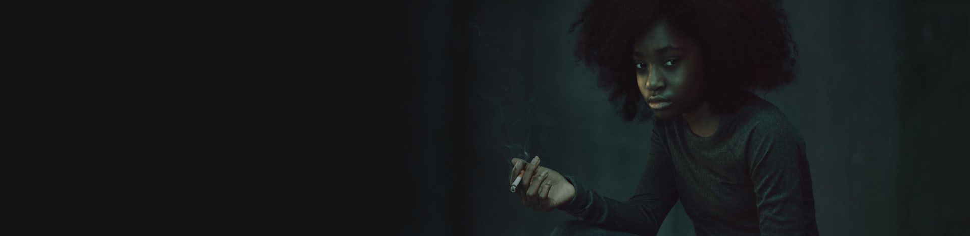 An African American/Black teen girl holding a cigarette in a dark room