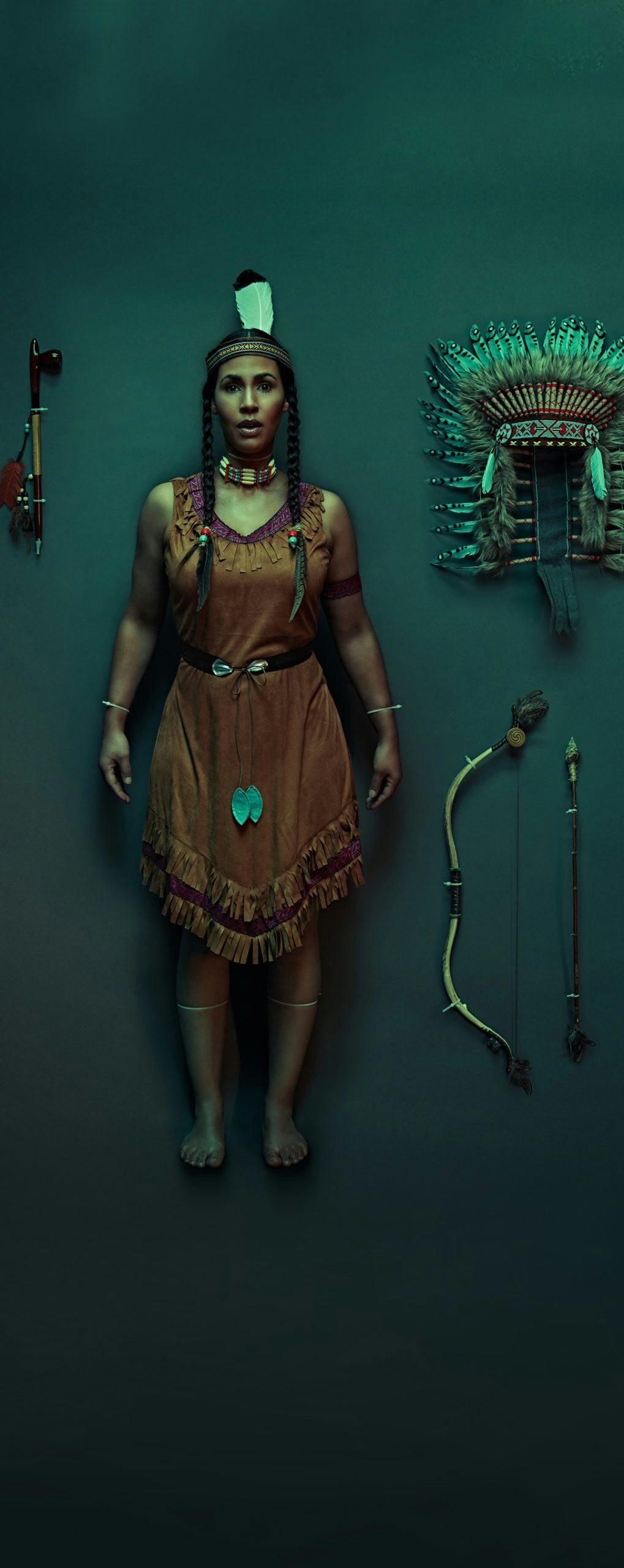 An American Indian woman dressed in traditional clothing standing in a dark room with American Indian accessories on display