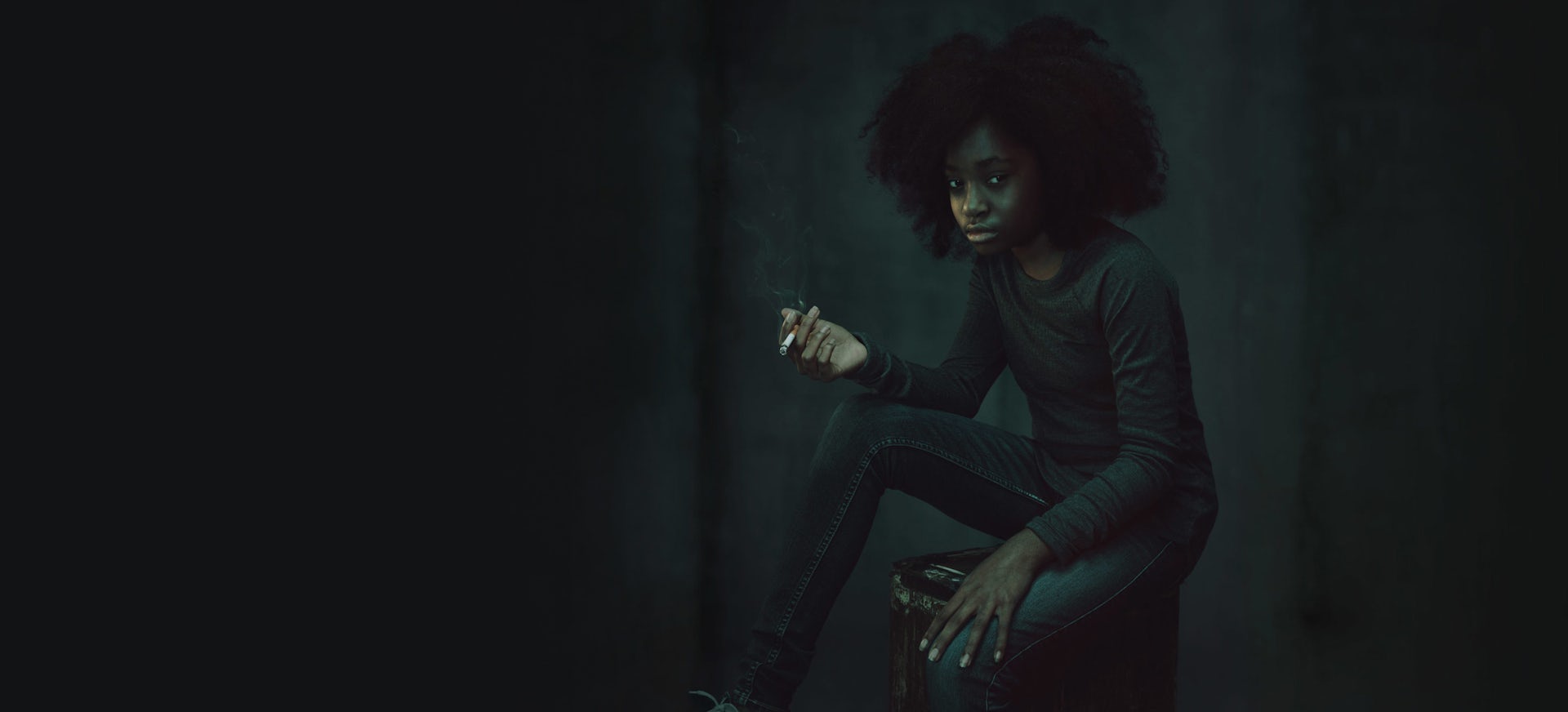 A young African American/Black teen girl sitting on a stool in a dark room holding a cigarette